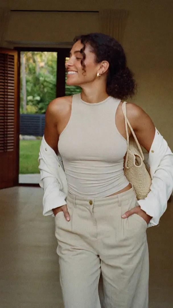 A woman with dark curly hair, in a sand top, open linen shirt and linen trousers, smiles as she steps through timber doors into a calm salon with white walls, shutters and boucle chairs.