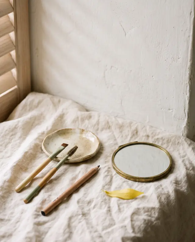 Spoolie brushes, a fine brow pencil, a small dish of brow tint and a brass hand mirror on cream linen by a lime-washed wall.